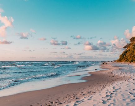Strand mit Sand und Wellen, rechts bewaldeter Küstenabschnitt unter blauem Himmel mit vereinzelten Wolken