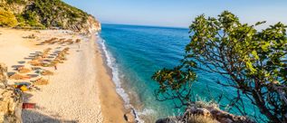 Blick auf den Gjipe Beach bei Himarë in Albanien mit hellem Sandstrand, türkisfarbenem Meer, Felsklippen und Sonnenschirmen entlang der Küste