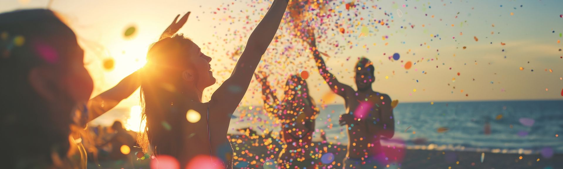 Mehrere Personen am Strand werfen buntes Konfetti in die Luft bei Sonnenuntergang über dem Meer.