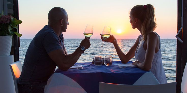 Ein Paar sitzt in einem Restaurant direkt am Meer bei Sonnenuntergang und halten Weingläser
