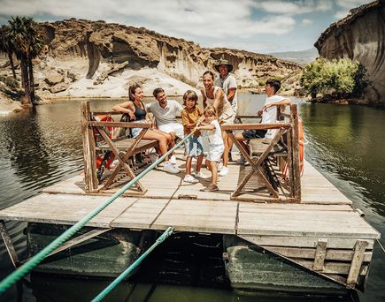 Mehrere Personen sitzen und stehen auf einem hölzernen Floß, das mit Seilen an Land befestigt ist, umgeben von felsiger Landschaft und Wasser auf Teneriffa.
