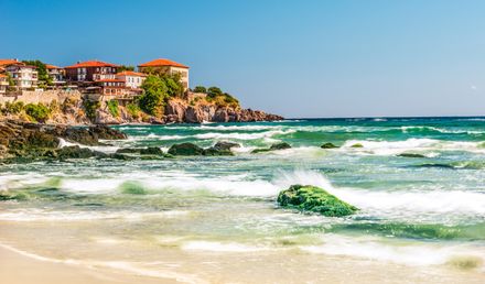 Küstenlandschaft in Bulgarien: Wellen treffen auf einen Sandstrand mit Felsen, im Hintergrund die Altstadt von Sosopol mit Häusern auf einer Klippe über der Bucht.