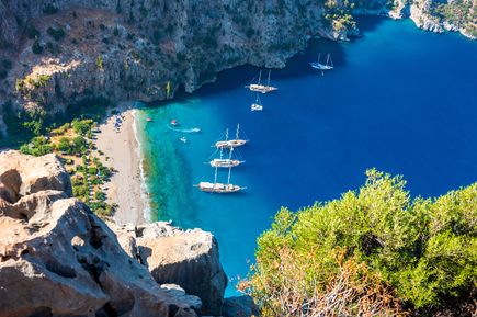 Steile Felswände und eine abgelegene Bucht mit klarem Blau, segelnden Booten und einem Naturstrand im türkischen Butterfly Valley, auch Schmetterlingstal genannt