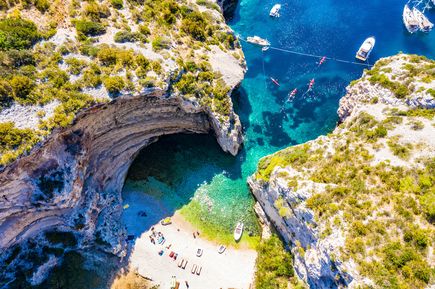 Luftaufnahme der Uvala Stiniva auf der Insel Vis in Kroatien mit steilen Felsklippen, kleinem Kiesstrand, türkisblauem Wasser sowie Booten und Kajaks in der geschützten Adria-Bucht