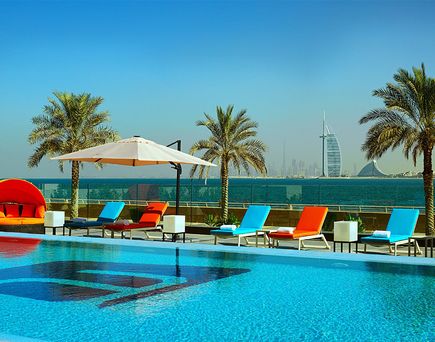 Poolbereich des Aloft Palm Jumeirah Hotels mit Liegestühlen in Blau und Orange, Sonnenschirm und Palmen, im Hintergrund das Meer und der Burj Al Arab in Dubai.