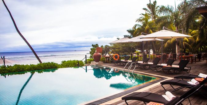 Pool des DoubleTree by Hilton Seychelles Allamanda Resort & Spa mit Sonnenliegen auf einer Holzterrasse, umgeben von Palmen und mit direktem Blick auf das Meer.