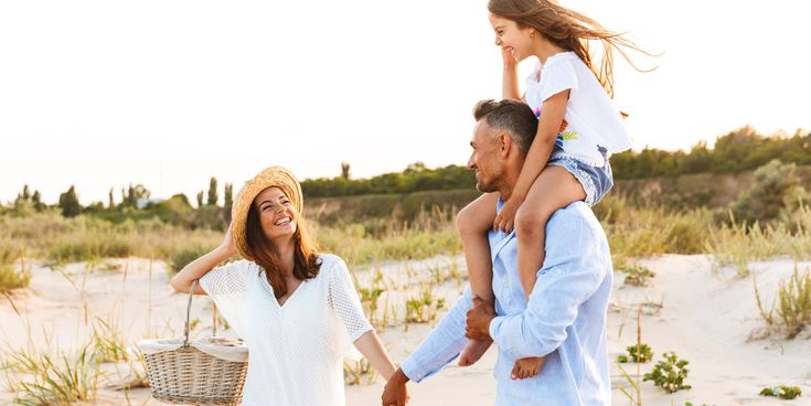 Familie spaziert an einem sonnigen Dünenstrand entlang; der Mann trägt das Kind auf den Schultern, während die Frau einen Picknickkorb hält. Hinter ihnen liegen Sand, Gräser und eine natürliche Küstenlandschaft