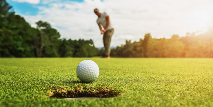 Golfball auf dem Grün vor einem Loch auf einem Golfplatz, während eine Person im Hintergrund einen Schlag ausführt