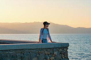 Frau mit Cap und offenem Hemd lehnt an einer Steinmauer mit Blick auf das Meer und Berge im Hintergrund