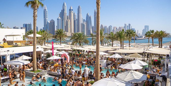 Blick auf belebten Poolbereich mit vielen Menschen, weißen Sonnenschirmen und Palmen, im Hintergrund die Wolkenkratzer von Dubai, aufgenommen im FIVE Palm Jumeirah Dubai.