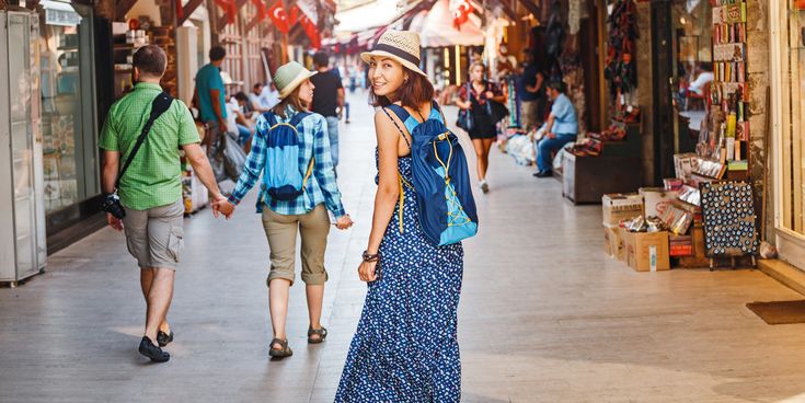 Touristin mit Rucksack und Sommerkleid geht durch eine belebte Marktgasse in der Türkei und blickt lächelnd zurück; umgeben von Geschäften und anderen Reisenden