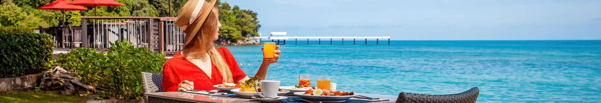 Frau in roter Kleidung sitzt an einem Frühstückstisch mit Kaffee, Saft und Speisen direkt am türkisblauen Meer und genießt den Meerblick