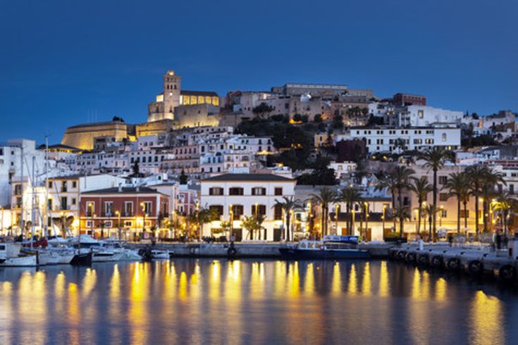 Blick auf die beleuchtete Altstadt von Ibiza-Stadt am Abend mit Hafen, Palmen und der Festung Dalt Vila auf einem Hügel