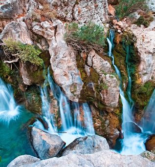 Mehrere schmale Wasserfälle fließen über moosbedeckte Felsen in ein türkisfarbenes Becken.