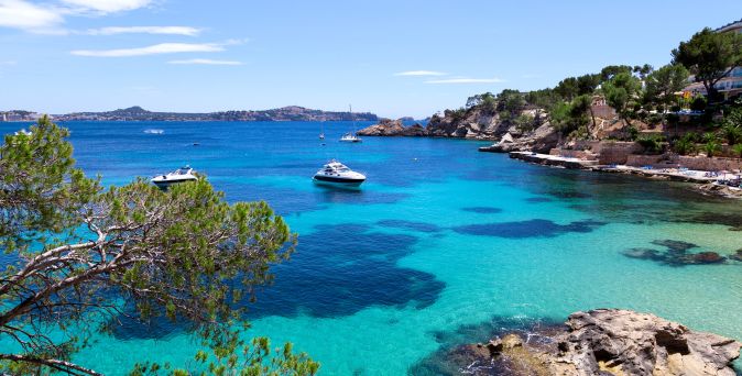 Blick auf das klare, türkisfarbene Wasser der Balearen mit zwei Booten im Vordergrund und felsigen Ufern im Hintergrund.