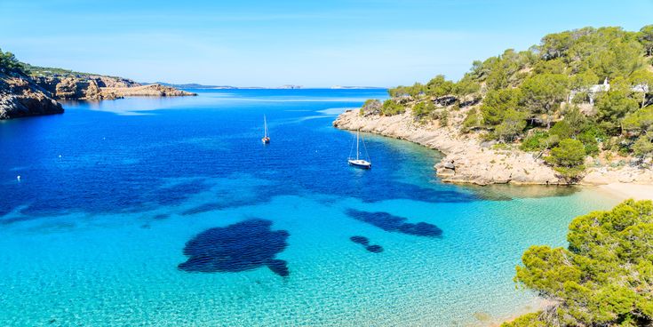 Bucht Cala Salada auf Ibiza mit klarem türkisblauem Wasser, Segelbooten und bewaldeten Felsen am Ufer unter blauem Himmel.