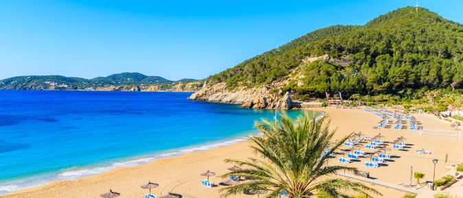 Panoramablick auf den Strand Cala de San Vicent auf Ibiza mit goldenem Sand, klarem blauen Wasser, Sonnenliegen und bewaldeten Hügeln im Hintergrund