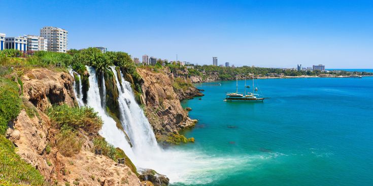 Düden-Wasserfall in Antalya stürzt von einer Felsklippe ins Mittelmeer, mit Booten auf blauem Wasser und Stadt im Hintergrund