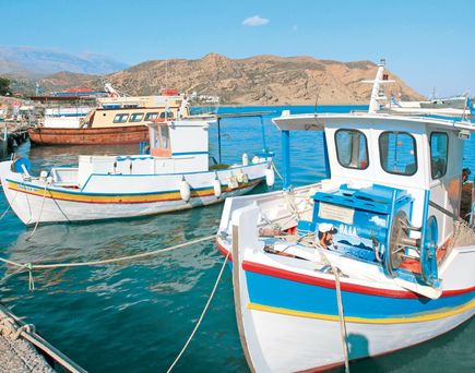 Fischerboote mit weißen Rümpfen und bunten Streifen liegen an einem Hafenpier vor einer felsigen Küstenlandschaft unter blauem Himmel.