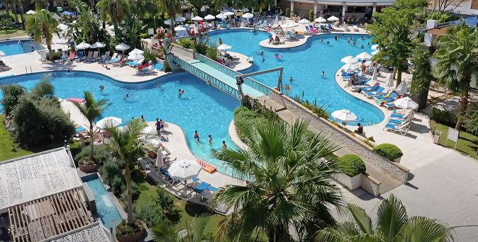 Großer Hotelpool mit Brücke, umgeben von Liegestühlen, Sonnenschirmen und Palmen