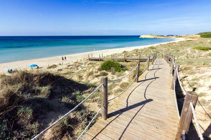 Holzsteg mit Seilen führt durch die Dünen zum langen Sandstrand von Son Bou auf Menorca, mit türkisblauem Meer im Hintergrund