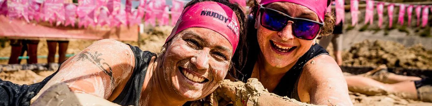 Zwei Teilnehmende beim Muddy Angel Run, mit Schlamm bedeckt an einem Hindernisparcours, tragen pinke Stirnbänder mit Event-Branding vor Fahnen der Veranstaltung