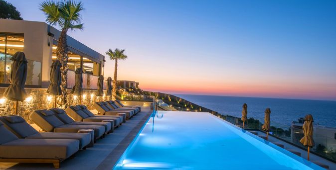 Beleuchteter Infinity-Pool mit Liegen und Palmen vor einem Hotel, Blick aufs Meer bei Sonnenuntergang.