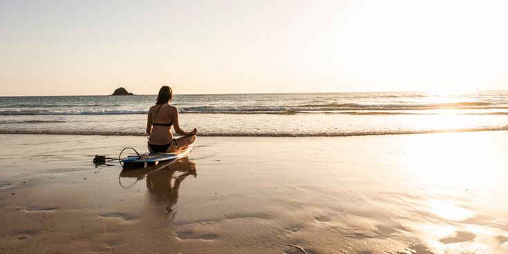 Frau im Bikini sitzt im Schneidersitz auf ihrem Surfbrett am Strand und blickt beim Sonnenuntergang aufs Meer