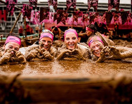 Vier Teilnehmerinnen mit pinken Stirnbändern liegen in schlammigem Wasser und halten ihre Hände zusammen in Herzform