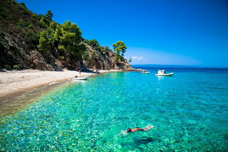 Klares türkisfarbenes Meer mit Schwimmer, felsige Küste mit Pinien und kleinen Booten im Wasser.