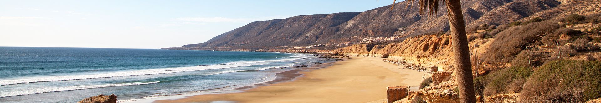 Panoramaansicht des Strandes von Taghazout in Marokko, mit sanften Wellen, goldenem Sand und bewaldeten Hügeln im Hintergrund.