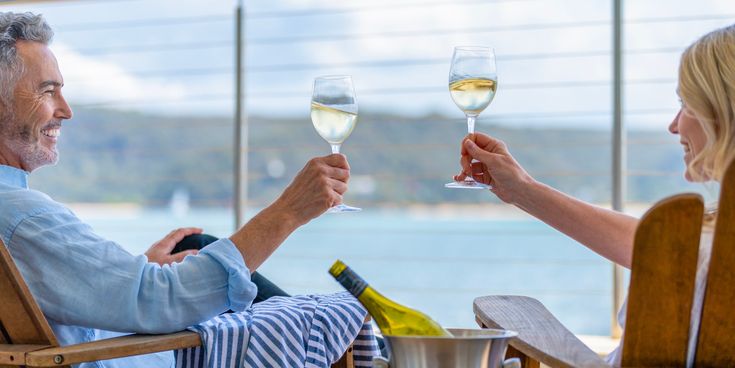 Mann und Frau sitzen entspannt auf einem Balkon mit Meerblick und stoßen mit Gläsern Weißwein an; auf einem kleinen Tisch steht eine offene Weinflasche.