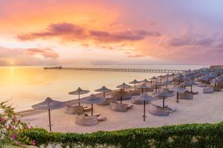 Strand mit Liegen und Sonnenschirmen bei Sonnenuntergang, langer Steg im ruhigen Meer in Marsa Alam