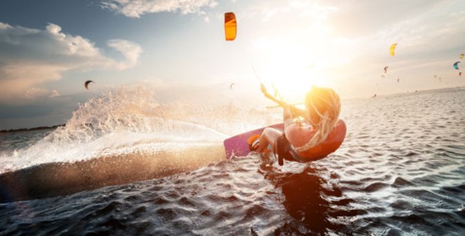 Kitesurfer gleitet dynamisch über das Wasser, während im Hintergrund mehrere Kites in der Luft schweben, Sonnenuntergangsstimmung