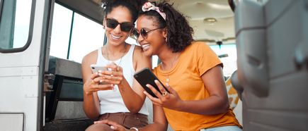 Zwei junge Frauen sitzen lachend im Camper und schauen gemeinsam auf ihre Smartphones