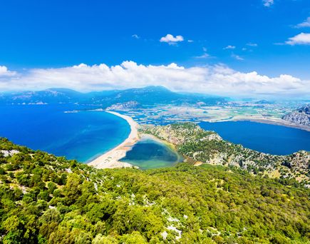 Luftaufnahme des Iztuzu-Strandes in der Türkei mit schmaler Sandbank zwischen zwei Lagunen, umgeben von grünen Hügeln und tiefblauem Meer unter sonnigem Himmel