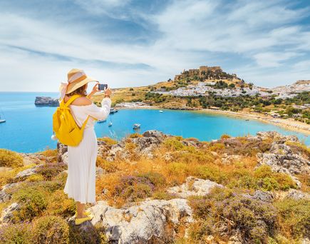 Frau mit gelbem Rucksack und Strohhut fotografiert die Bucht von Lindos mit Burg auf dem Hügel und Segelboot im Meer.