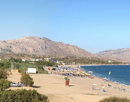 Strand mit Sonnenschirmen und Liegen, dahinter Hügel und klares blaues Meer unter hellem Himmel.