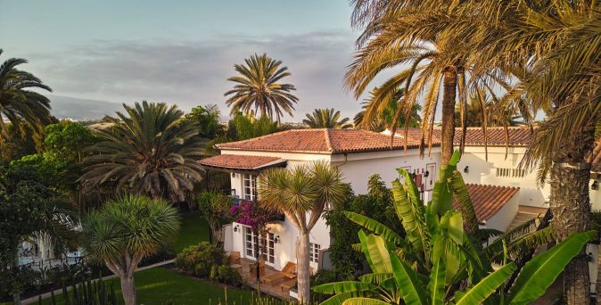 Zwei-stöckiges Hotelgebäude mit Terrakottadach, umgeben von Palmen und tropischen Pflanzen. Blühende Pflanzen sind an der Fassade sichtbar.