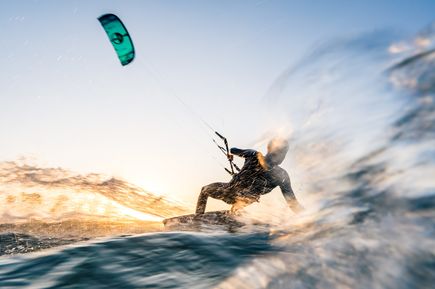 Ein Windsurfer in einem schwarzen Neoprenanzug reitet auf einer Welle, während ein Kiteschirm in Blau und Schwarz über ihm schwebt