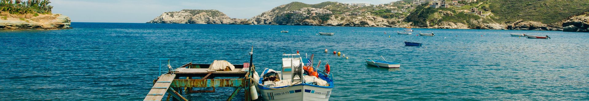 Blick auf einen kleinen Hafen mit mehreren Booten auf ruhigem Wasser, umgeben von felsigen Küsten und bewachsenen Hügeln.