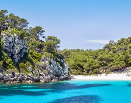 Klares türkisfarbenes Wasser in einer Bucht umgeben von steilen Felsen und grünen Kiefern auf Menorca