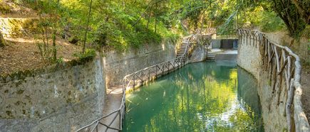 Schmaler Wasserkanal mit steinernen Ufern und Holzgeländern, umgeben von dichtem Grün und Bäumen.