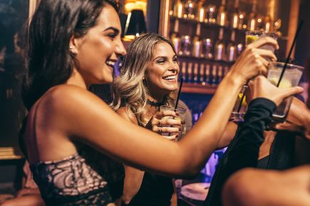 Zwei junge Frauen stoßen lachend mit Cocktails in einer Bar an, im Hintergrund Regale mit Flaschen