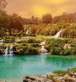 Mehrstufiger Wasserfall fließt in türkisfarbenen See, umgeben von grünen Bäumen und Hügeln bei Sonnenuntergang