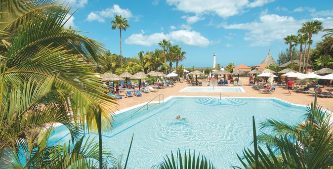 Weitläufige Poollandschaft des IFA Altamarena by Lopesan Hotels auf Fuerteventura, umgeben von Palmen, Sonnenschirmen und Liegen; im Hintergrund ist der Leuchtturm von Morro Jable vor blauem Himmel zu sehen