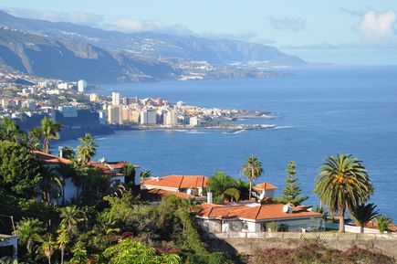 Blick auf eine Küstenstadt mit roten Dächern, Palmen und Bergen im Hintergrund am Meer