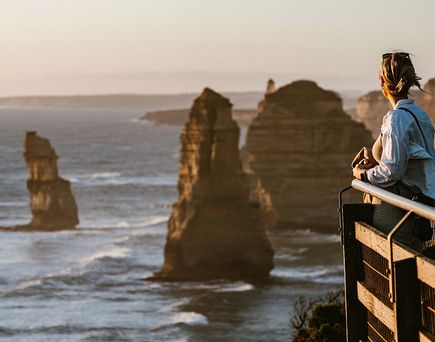 Person mit hellem Hemd blickt von Aussichtsplattform auf Felsformationen im Meer bei Sonnenuntergang.