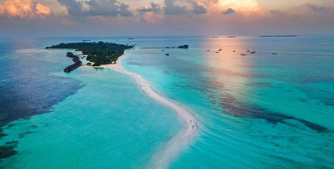 Luftaufnahme einer schmalen Sandbank, die zu einer Insel mit Vegetation und Gebäuden im türkisfarbenen Meer führt, unter einem bewölkten Himmel bei Sonnenuntergang