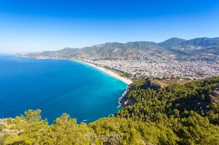 Blick auf den Kleopatra-Strand in Alanya mit langem Sandstrand, blauem Meer, Stadt und bewaldeten Hügeln im Hintergrund.